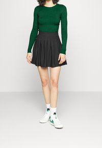 Maglietta verde a righe a maniche lunghe, gonna nera plissettata, sneakers bianche con accenti verdi e calzini bianchi. Focus sugli elementi di un outfit casual e a strati.