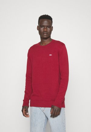 Suéter rojo de punto con cuello redondo, puños y bajo acanalados. Presenta un pequeño logo en el pecho, combinado con unos jeans de color azul claro.