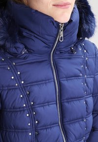 Tmavě modrá nafukovací bunda s vysokým límcem, stříbrným zipem a ozdobnými stříbrnými nýty. Má lem kapuce z umělé kožešiny a prošívaný vzor.