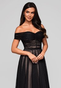 Robe de soirée noire à épaules dénudées avec un corsage froncé et une jupe transparente à pois. Comprend une taille ajustée et un léger éclat dans le tissu.