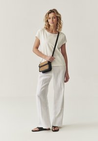 T-shirt beige clair à manches courtes, pantalon long en lin blanc, sandales noires et un petit sac bandoulière noir orné de détails dorés.