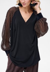 Czarna bluzka z dekoltem w serek i teksturowanymi bufkami z przezroczystej brązowej siateczki w kratkę. Luzny fason z układającą się tkaniną.
