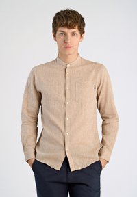 Camicia button-up marrone chiaro, con texture, colletto a band, taschino frontale e bottoni bianchi. Indossata con pantaloni blu navy, mani in tasca.