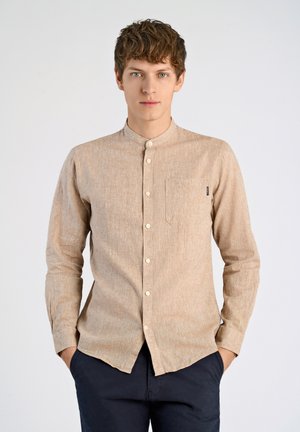 Camisa de botón texturizada de color marrón claro con cuello tipo banda, bolsillo frontal y botones blancos. Usada con pantalones azul marino, con las manos en los bolsillos.