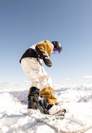 Un snowboardeur casqué, vêtu d’une tenue jaune et blanche, trace un virage dans la neige sur une montagne, avec des sommets enneigés sous un ciel bleu dégagé.