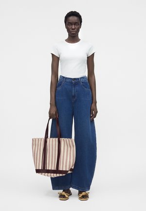 Γυναίκα με λευκό εφαρμοστό t-shirt και φαρδιά μπλε τζιν παντελόνια, κρατώντας μπεζ και καφέ ριγέ τσάντα, στέκεται μπροστά σε απλό φόντο.