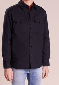 Camicia blu scuro con bottoni neri, due tasche sul petto, colletto e maniche lunghe. Indossata sopra una camicia a scacchi rossi e neri.