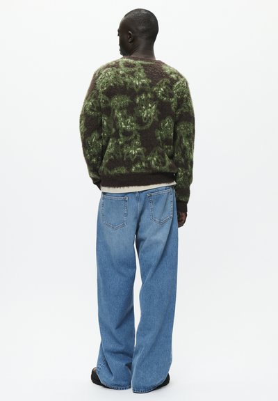 Grønn og brun fluffy genser med abstrakt mønster, sammen med lyseblå, løstsittende jeans. Modell vist fra bak.