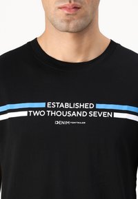 Svart t-skjorte med korte ermer, rund halsutskjæring og trykket tekst i hvite og blå striper, med "DENIM TOM TAILOR" nederst.
