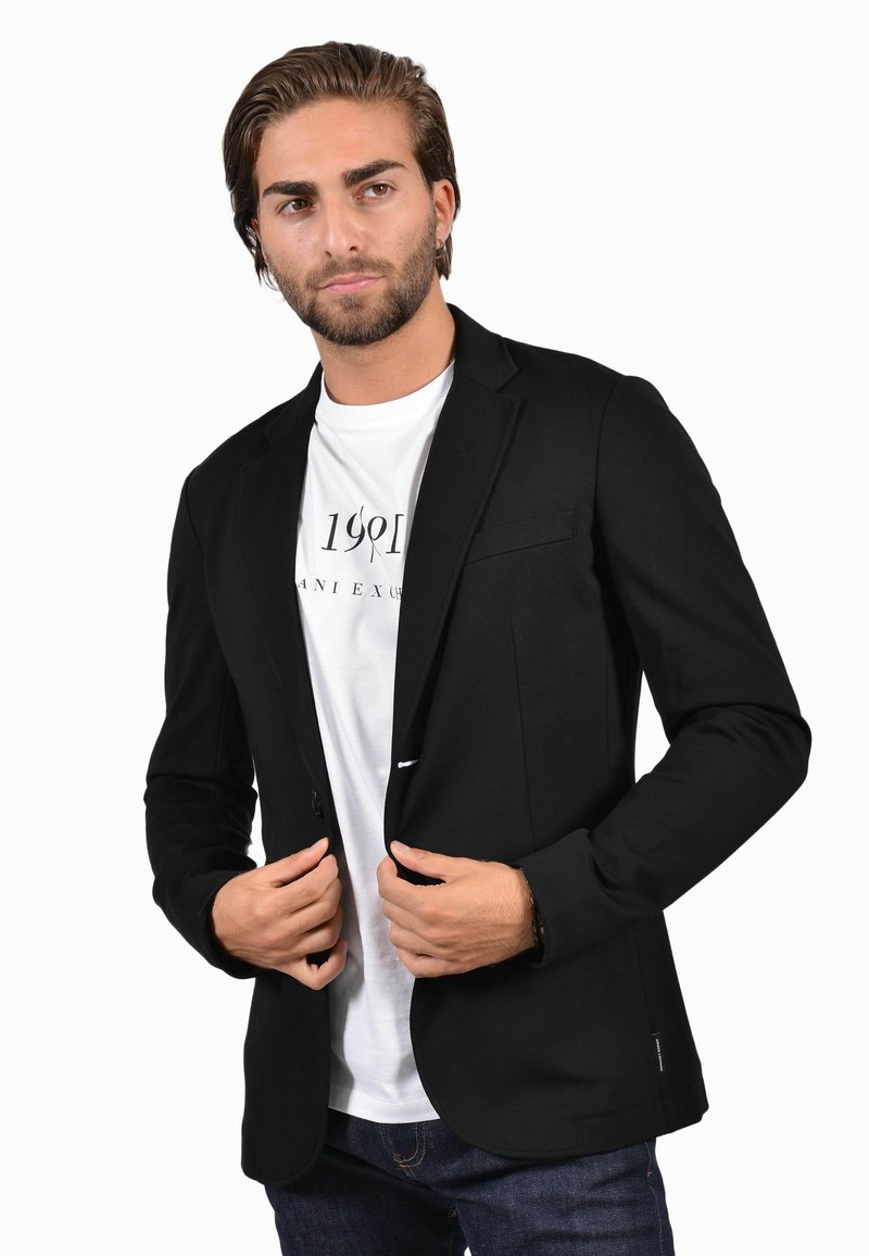 Blazer nero con chiusura a un bottone, vestibilità slim e rever a punta, indossato sopra una t-shirt bianca con grafica e jeans scuri.