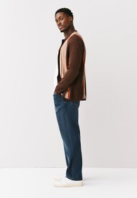 Uomo in piedi di lato con le mani in tasca, indossa un cardigan marrone a righe, jeans blu, sneaker bianche, guarda la fotocamera su uno sfondo semplice.