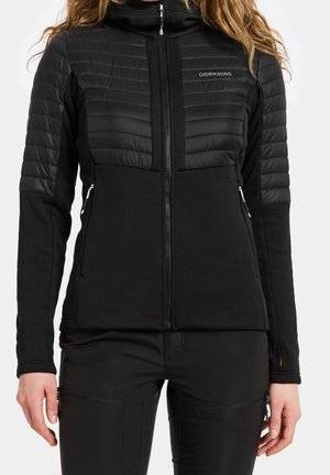 Woman wearing a black zip-up jacket with padded shoulders and arms, featuring side zipper pockets and thumbholes on the sleeves.