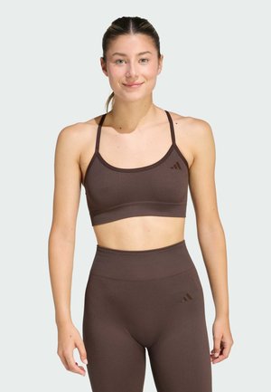 Junge Frau trägt braunen nahtlosen Sport-BH und hoch taillierte Leggings, steht und lächelt vor einem schlichten hellen Hintergrund.