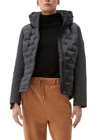 Gepolsterte schwarze Jacke mit Kapuze, die eine gesteppten Vorderseite und glatte Ärmel hat. Kombiniert mit braunen Jogginghosen mit Kordelzug.