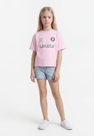 Νεαρή κοπέλα όρθια, φοράει ροζ μπλουζάκι με την επιγραφή «LEAGUE», ανοιχτόχρωμα τζιν σορτς και λευκά αθλητικά παπούτσια σε λευκό μονόχρωμο φόντο.