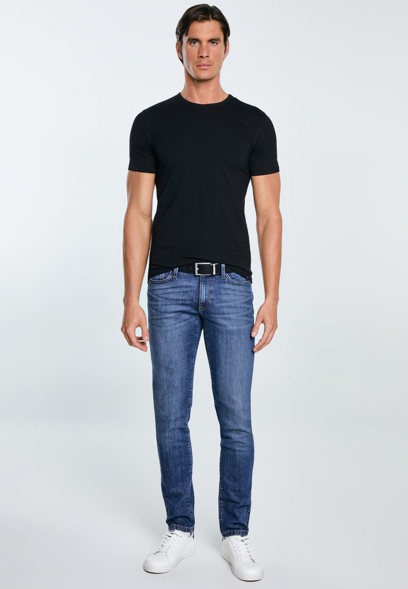 Mees seisab eespool, kannab musta T-särki, siniseid teksapükse musta vööga ja valgeid tossusid heleda ühevärvilise tausta ees.