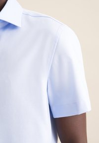 Camicetta azzurra a maniche corte con colletto classico, texture liscia e cuciture pulite lungo le cuciture delle spalle.