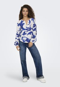 Weißer Pullover mit blauem Blumenmuster, V-Ausschnitt, langen Ärmeln, kombiniert mit weiten Denim-Jeans und weißen Sneakern. Schlicher Hintergrund.