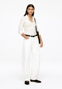 Woman in white ribbed long-sleeve shirt and wide-leg white pants with black belt, black shoes, and shoulder bag stands against white background.