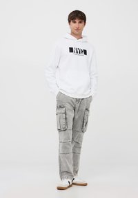 Felpa bianca con cappuccio e scritta "NYC Times Square", abbinata a pantaloni cargo grigi chiari con più tasche e una texture sbiadita.