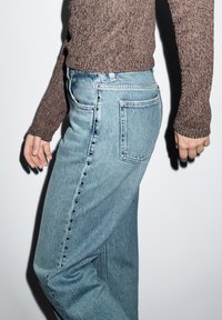 Světle modré džíny s vysokým pasem, s černými patentními knoflíky na straně a mírně volným střihem. Hnědý texturovaný svetr.