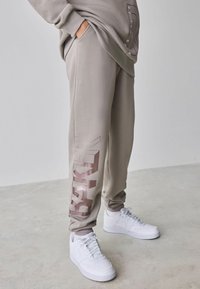 Lysegrå joggingbukser med glat tekstur, med en skinnende bronze logo på venstre ben. Kombineret med hvide sneakers.