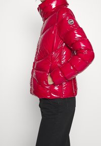 Röd glänsande puffjacka med quiltad design, hög krage och sidofickor. Har en cirkulär logotyp på vänster ärm.