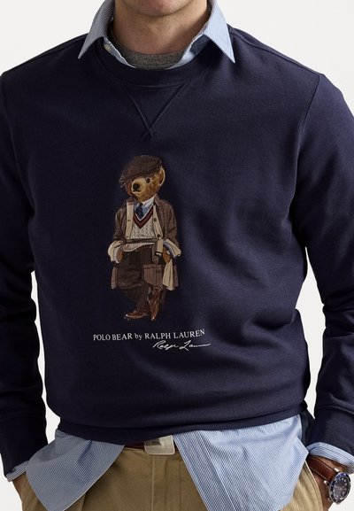 Sweatshirt marine avec un ours imprimé en tenue ajustée. Matériau en coton, col rond et poignets côtelés. Superposé à une chemise rayée.