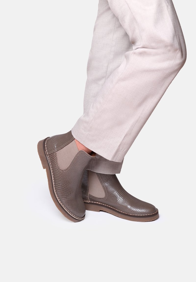 Bottines en cuir gris texturé avec panneaux latéraux élastiques, semelle contrastante et motif subtil de peau de serpent, associées à un pantalon de couleur claire.