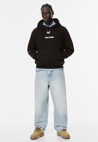 Ung person iført sort STWD STUDIO hættetrøje, blå stribet skjorte, lyse løse jeans og brune støvler, står med hænderne i hættetrøje lommen på hvid baggrund.