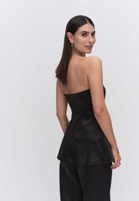 Musta olkaimeton satiinitoppi, jossa on istuva rinnanympärys ja levenevä helma, yhdistettynä laskeutuviin mustiin housuihin. Sujuva pinta ja minimaalinen muotoilu.