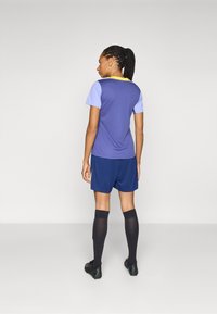 Lilla treningsskjorte med lyse lilla ermer, kombinert med marineblå shorts og svarte knesokker, med gule detaljer på shortsene.