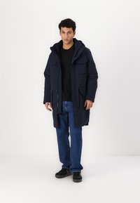 Parka bleu marine avec plusieurs poches avant, fermeture éclair et pressions, ainsi qu'une capuche, associée à un jean noir et des chaussures noires.