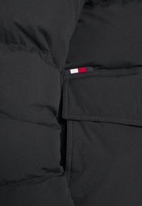 Czarna kurtka puchowa o gładkiej fakturze, z pionową kieszenią na zamek podkreślającą mały akcent w kolorach czerwonym, białym i niebieskim na tkaninie.