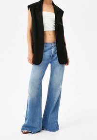 Svart ärmlös kavaj över en vit ribbad crop top, kombinerad med ljusblå vida jeans med slitna detaljer och fransad fåll.