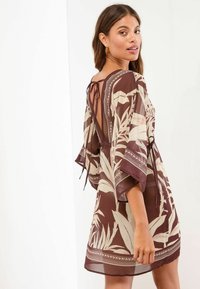 Abito kimono in marrone e beige chiaro, caratterizzato da un motivo floreale, maniche fluttuanti e un dettaglio con lacci sulla schiena per una vestibilità regolabile.