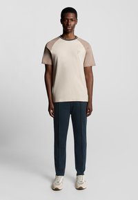 Beige und tannfarbene kurzärmelige T-Shirts mit runder, schwarz eingefaßter Ausschnitt, kombiniert mit dunkelblauen Hosen und cremefarbenen Turnschuhen.