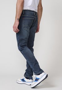 Jeans de mezclilla azul de corte ajustado con bolsillos traseros, detalles de costura sutil y una textura suave. Combinados con zapatillas deportivas blancas y azules.