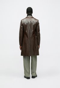 Bruine leren jas met een glanzende afwerking, lange mouwen en een getailleerde pasvorm, gecombineerd met losse groene broek en zwarte schoenen.