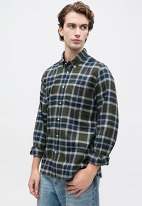 Fiatal férfi zöld és tengerészkék kockás ingben, felhajtott ujjakkal és világoskék farmernadrággal, egy sima fehér háttér előtt áll.