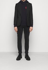 Blazer negro sobre una sudadera azul marino con un logo rojo, combinado con jeans oscuros y botas negras de cordones. La textura es suave y de corte ajustado.