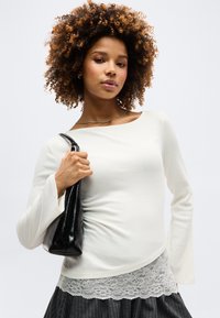 Γυναίκα με σγουρά μαλλιά που φοράει μπλούζα μακριάς μανίκας εκρού, εσώρουχο με δαντέλα, φούστα με ρίγες και κρατάει μια μαύρη τσάντα στον ώμο της.