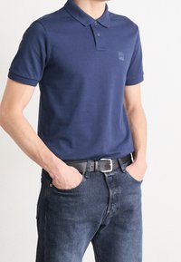 Námořnická modrá polo košile s límečkem, dvěma knoflíky a malým logem na prsou, spojená s tmavými džíny a černým opaskem.