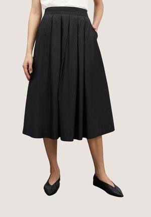 Vrouw die een zwarte geplooide midirok met zakken draagt en zwarte puntige platte schoenen, gecombineerd met een witte top, staand tegen een neutrale achtergrond.