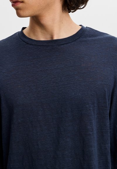 Jeune personne aux cheveux courts et foncés portant un t-shirt bleu marine uni à manches longues et col rond, tournée vers la gauche, sur un fond uni.