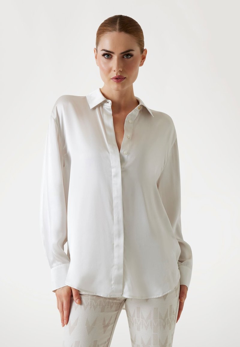 Femme aux cheveux tirés en arrière, portant une chemise blanche ample boutonnée et un pantalon clair à motif, sur un fond uni.