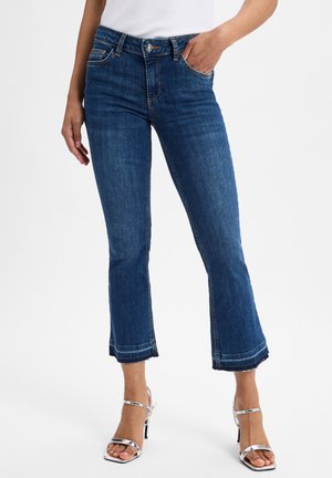 Femme portant un jean court bleu avec des ourlets effilochés et des sandales argentées à talons hauts, debout devant un fond uni.