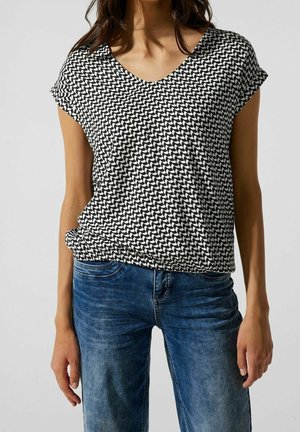 Vrouw draagt een zwart-wit geometrisch patroon top met korte mouwen, gecombineerd met een blauwe spijkerbroek, staand tegen een effen achtergrond.