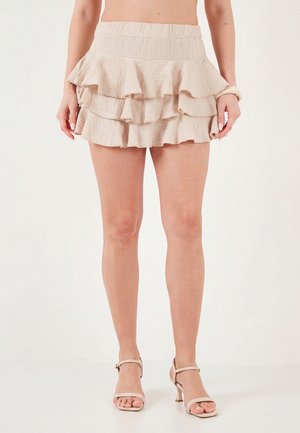 Žena v béžové minisukni s volánky v patrech a béžových sandálech na podpatku stojí na jednoduchém bílém pozadí.