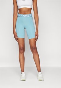 Kobieta stoi w jasnoniebieskich szortach rowerowych Nike Pro, białym staniku sportowym i białych butach sportowych na jednolitym tle.